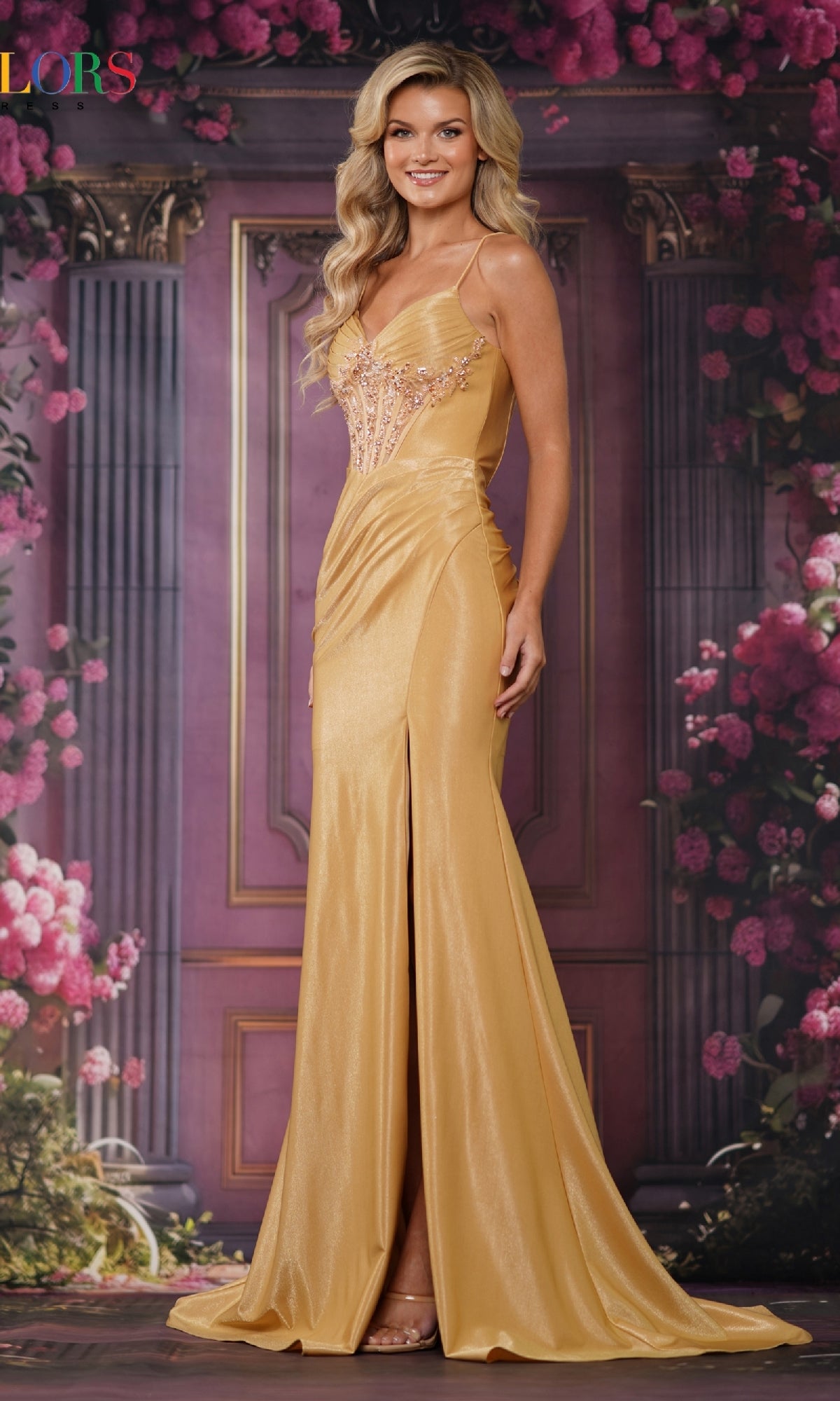 A woman smiles at the camera in front of a floral, columned backdrop, wearing the gold Colors Dress 3704—an elegant, floor-length sleeveless long prom dress.